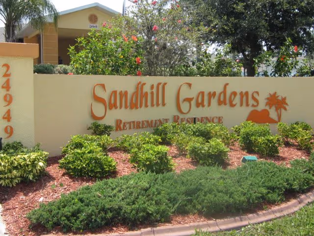 Exterior view of Sandhill Gardens Retirement Residence sign with landscaping and a building facade.