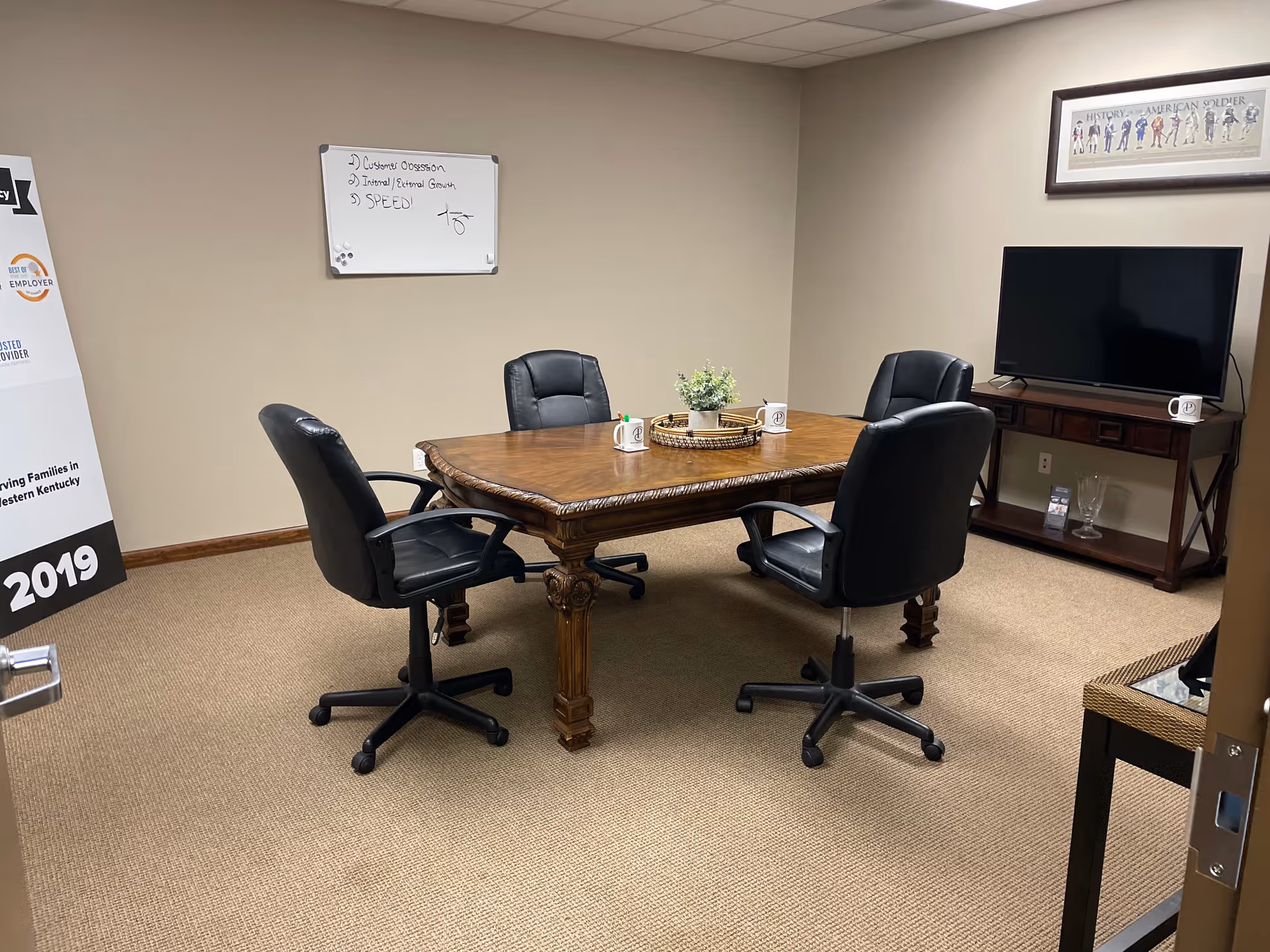 Beige conference room with wooden table, four black chairs, whiteboard, and a TV.