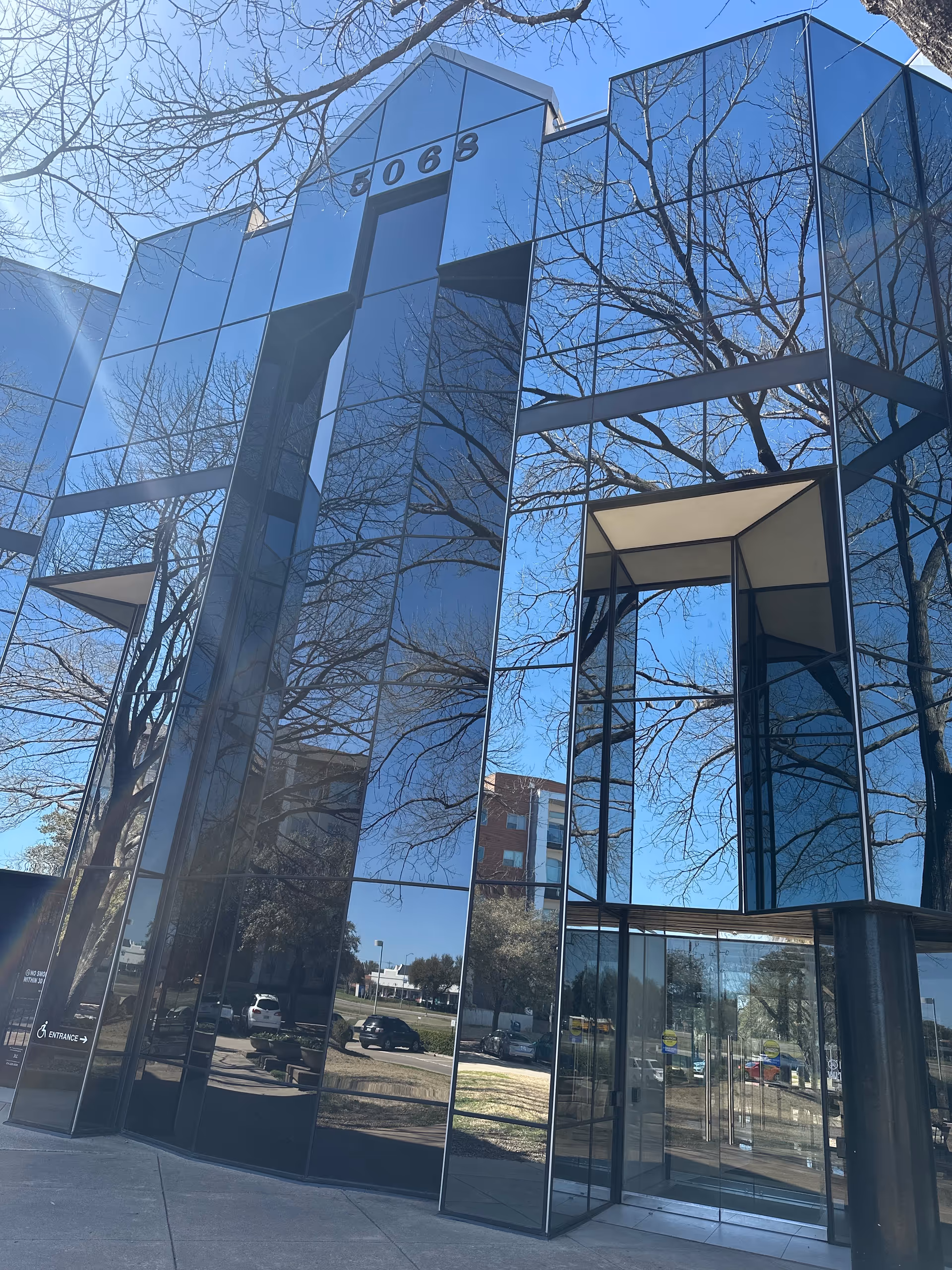 Modern glass-front building exterior reflecting bare trees and sky.