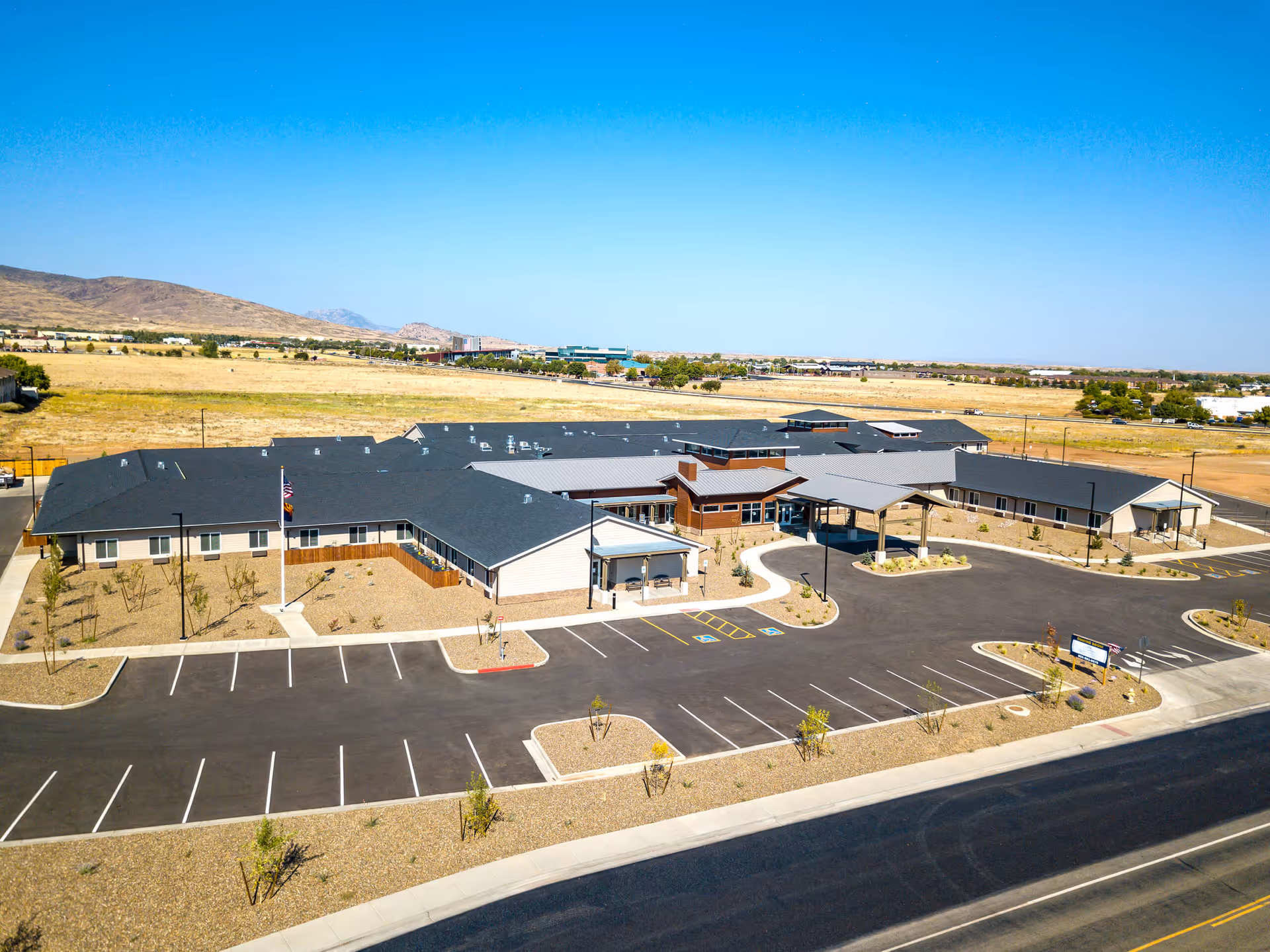 Aerial view of a single‑story senior living building with a covered main entrance and surrounding empty parking lot in a dry landscape.