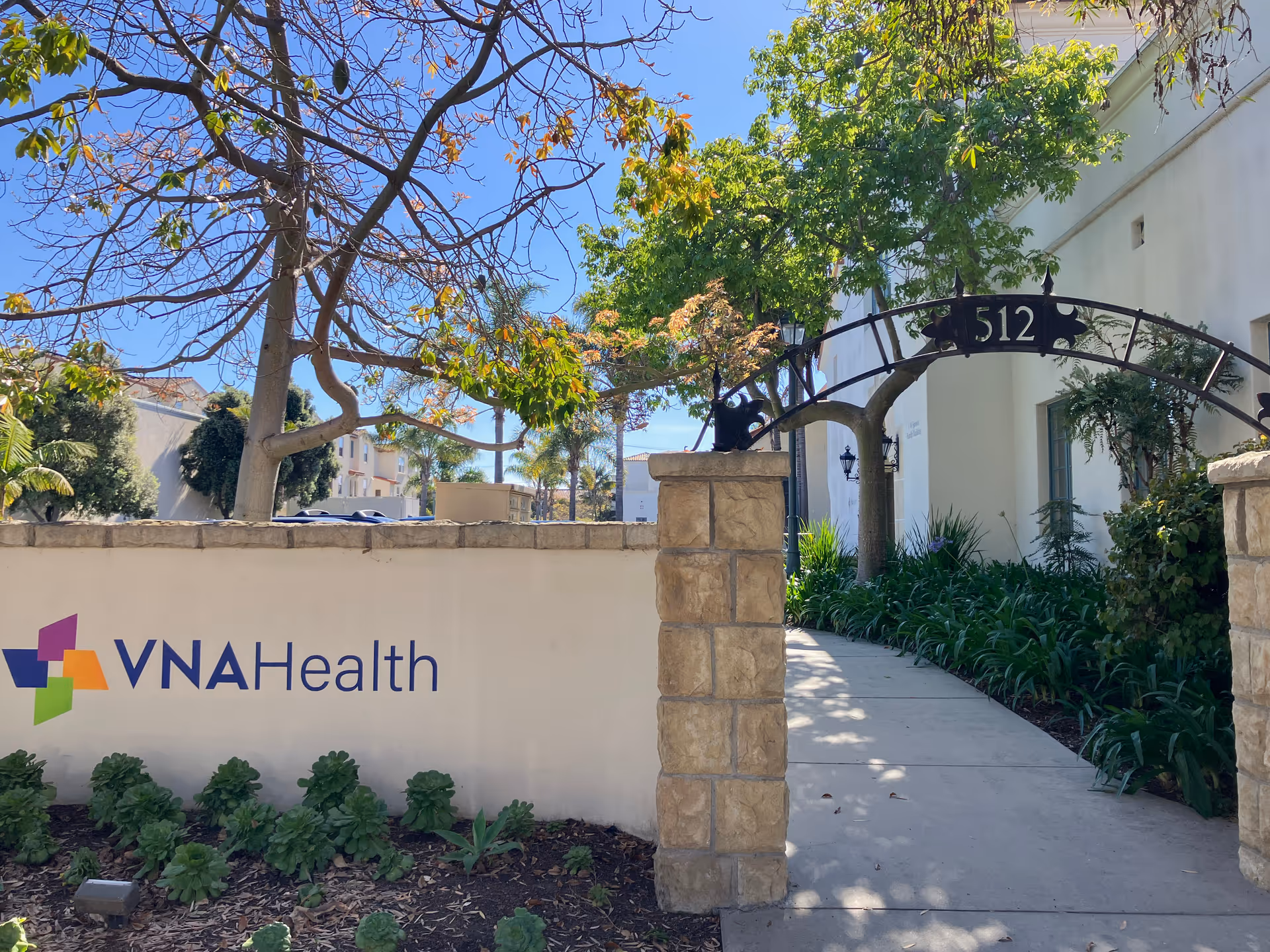 Exterior entrance to VNA Health facility with archway, walkway, and greenery.