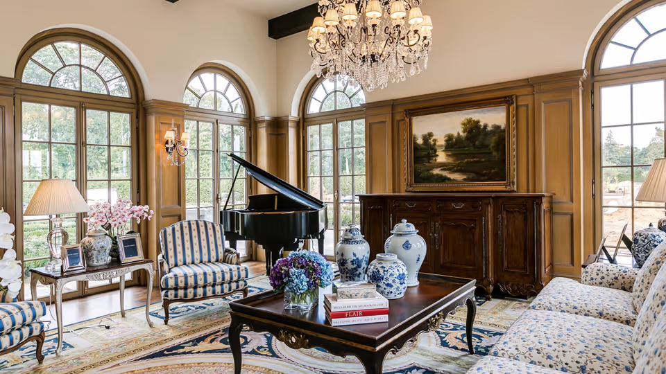 Elegant living room with a grand piano, chandelier, wood paneling, and blue upholstered seating.