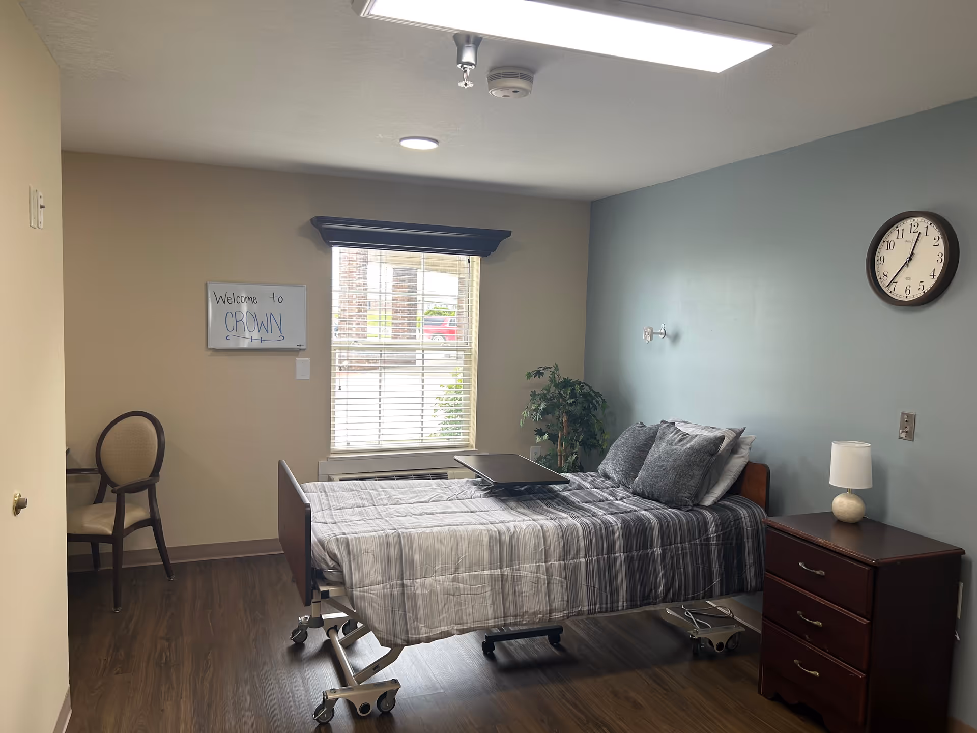 A hospital-style patient bedroom featuring a bed, nightstand, clock, chair, and window.
