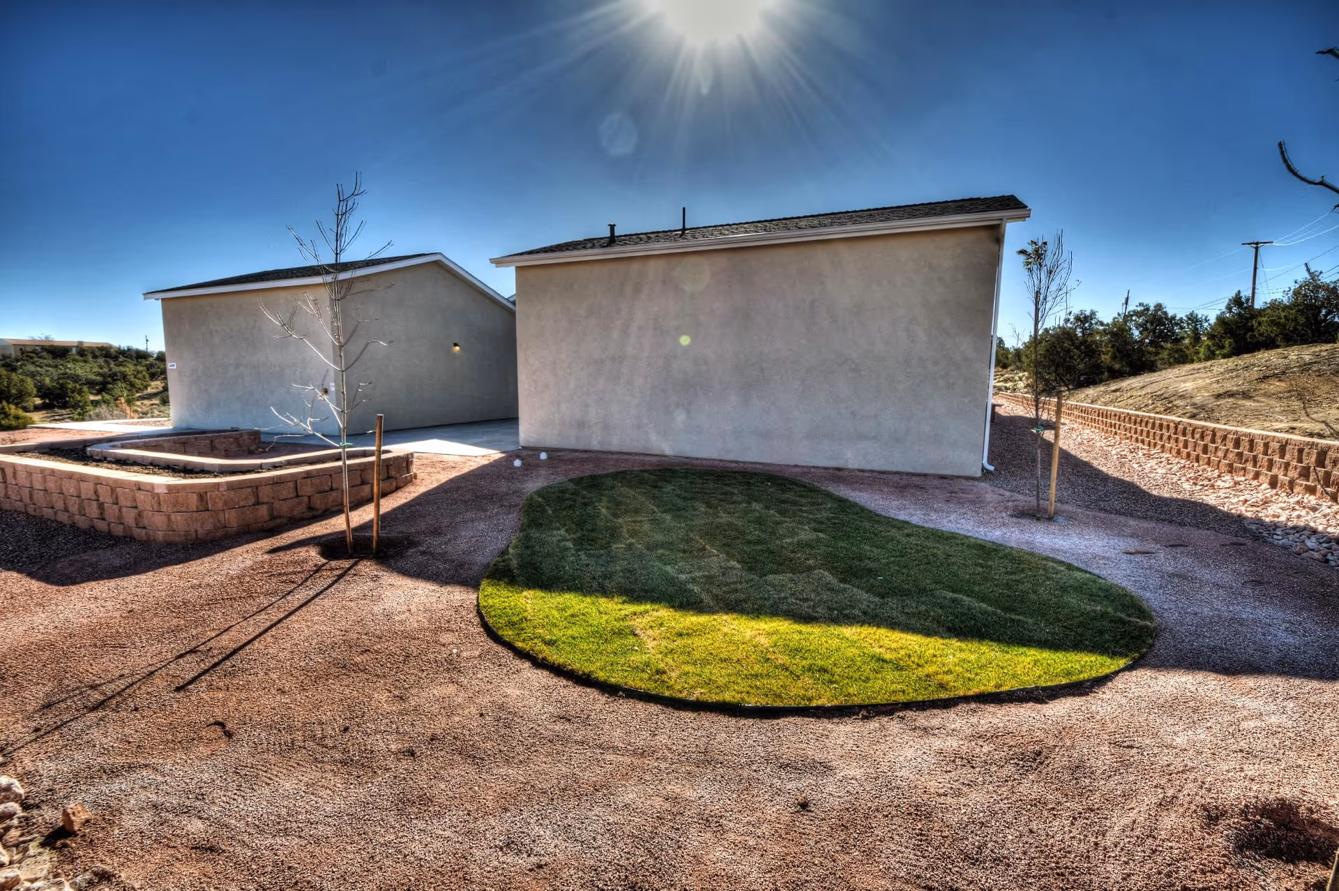 Outdoor view of a senior living facility showing two single-story buildings with beige walls and slanted roofs. The area between the buildings features a small patch of green grass surrounded by gravel and a few young trees. The sky is clear and sunny.