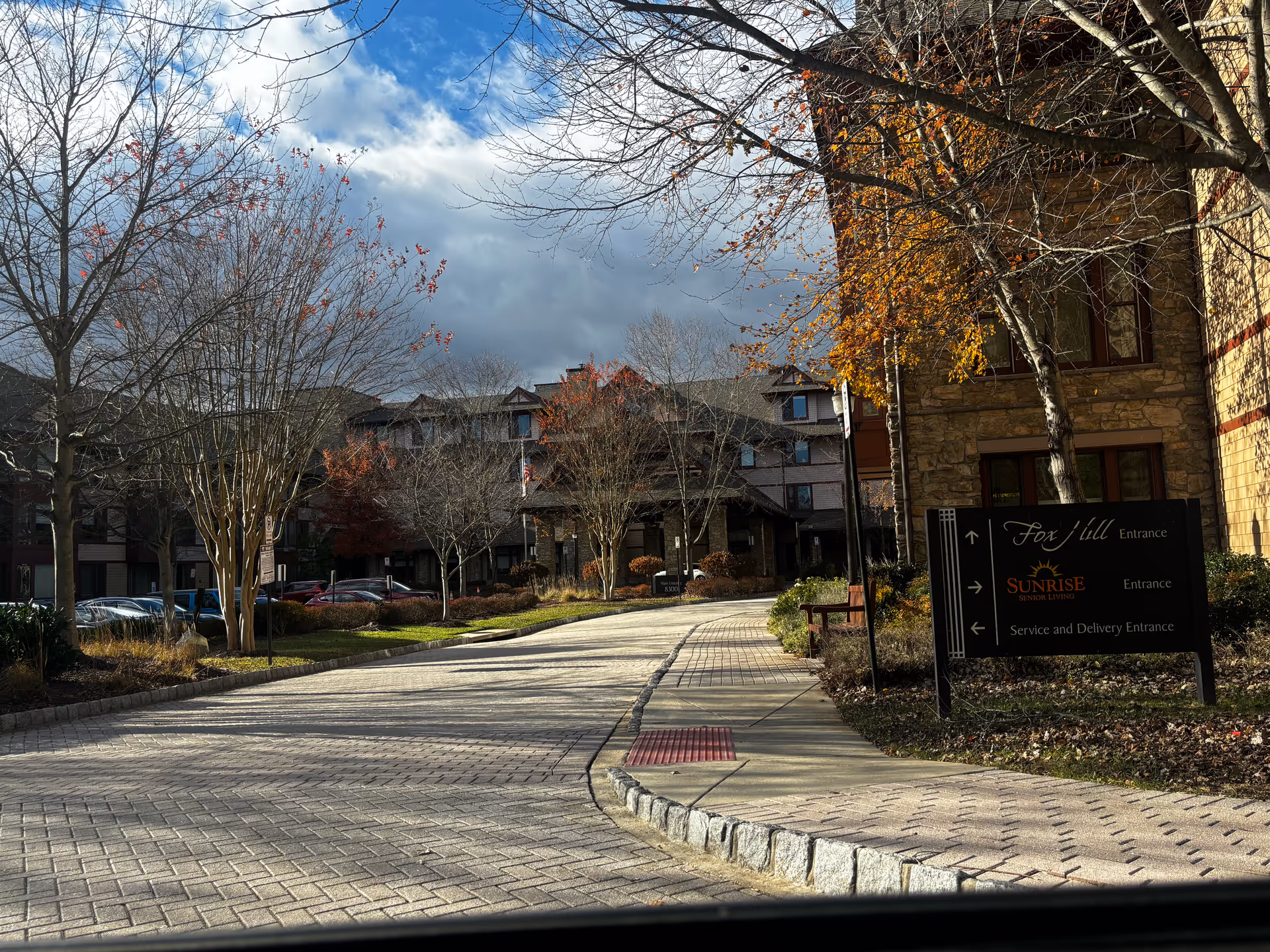 Driveway leading to the entrance of a senior living facility named Sunrise at Fox Hill, with a sign indicating directions for Fox Hill Entrance, Sunrise Senior Living Entrance, and Service and Delivery Entrance. The scene includes leafless trees, some with autumn leaves, parked cars, and a partly cloudy sky.