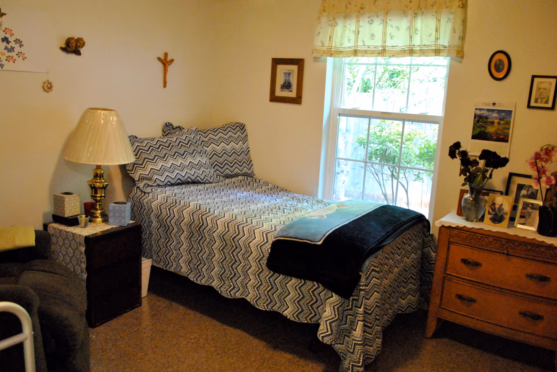 A small bedroom with a twin bed dressed in chevron-patterned bedding, a lamp on a nightstand, and a dresser with framed photos beside a window.