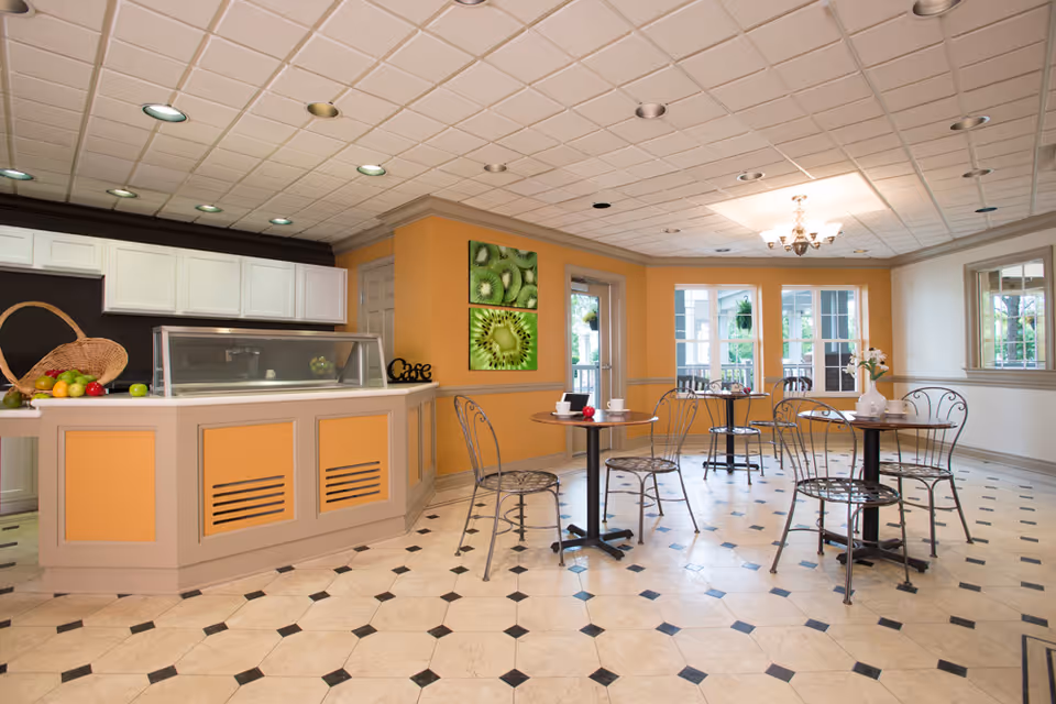 A bright dining area with several small round tables and metal chairs. The room has a tiled floor with a black and white pattern, orange and beige walls, and a ceiling with recessed lighting and a chandelier. There is a counter with a glass display case and a basket of fruit on it. Two framed pictures of kiwi fruit hang on the wall near a door leading outside to a patio area with more seating.