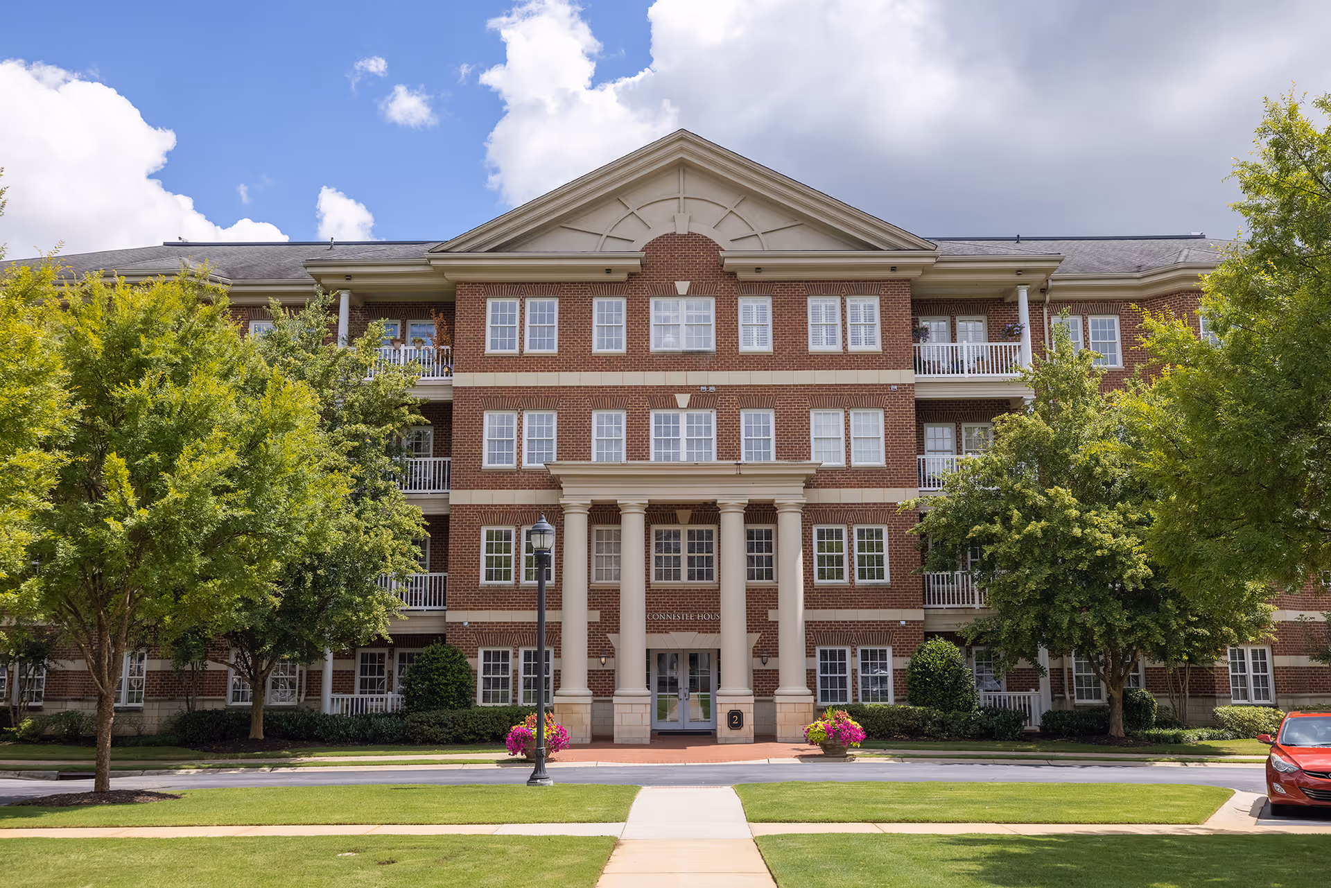 Front exterior view of a three-story brick building with white columns at the entrance, surrounded by green trees and a well-maintained lawn under a partly cloudy sky.