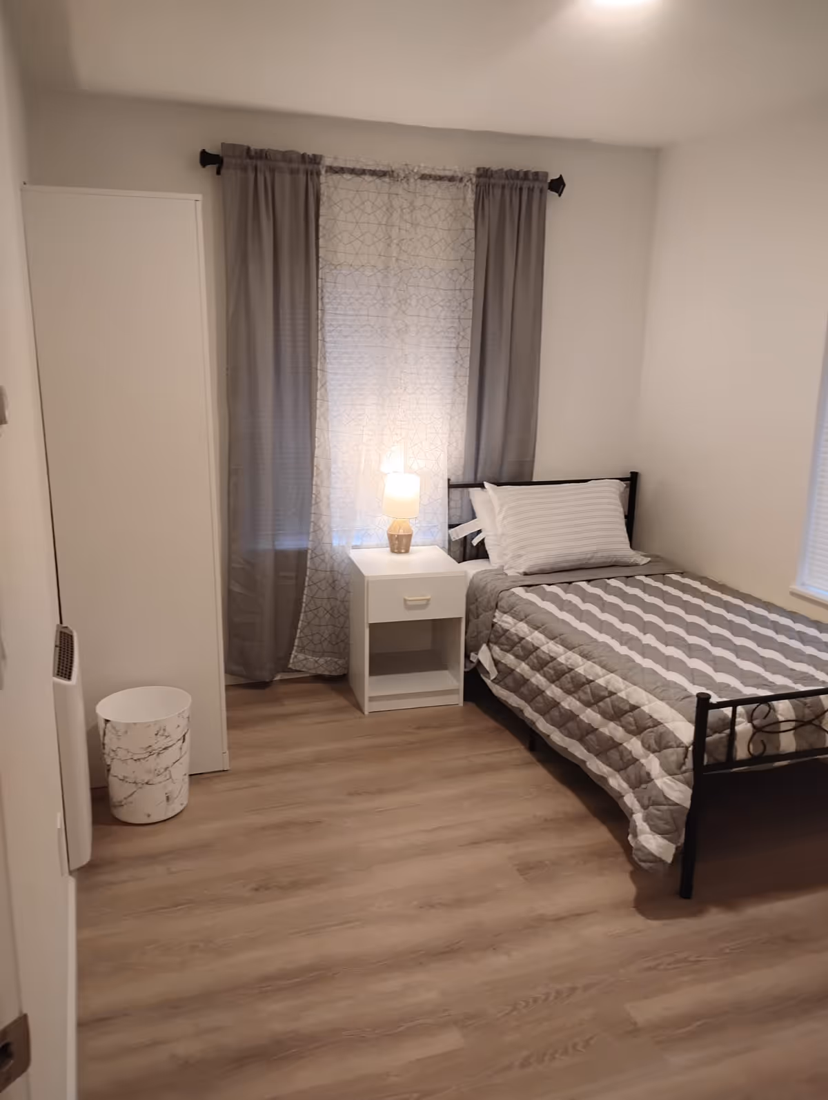 A tidy bedroom with a single bed, nightstand, lamp, and gray curtains.