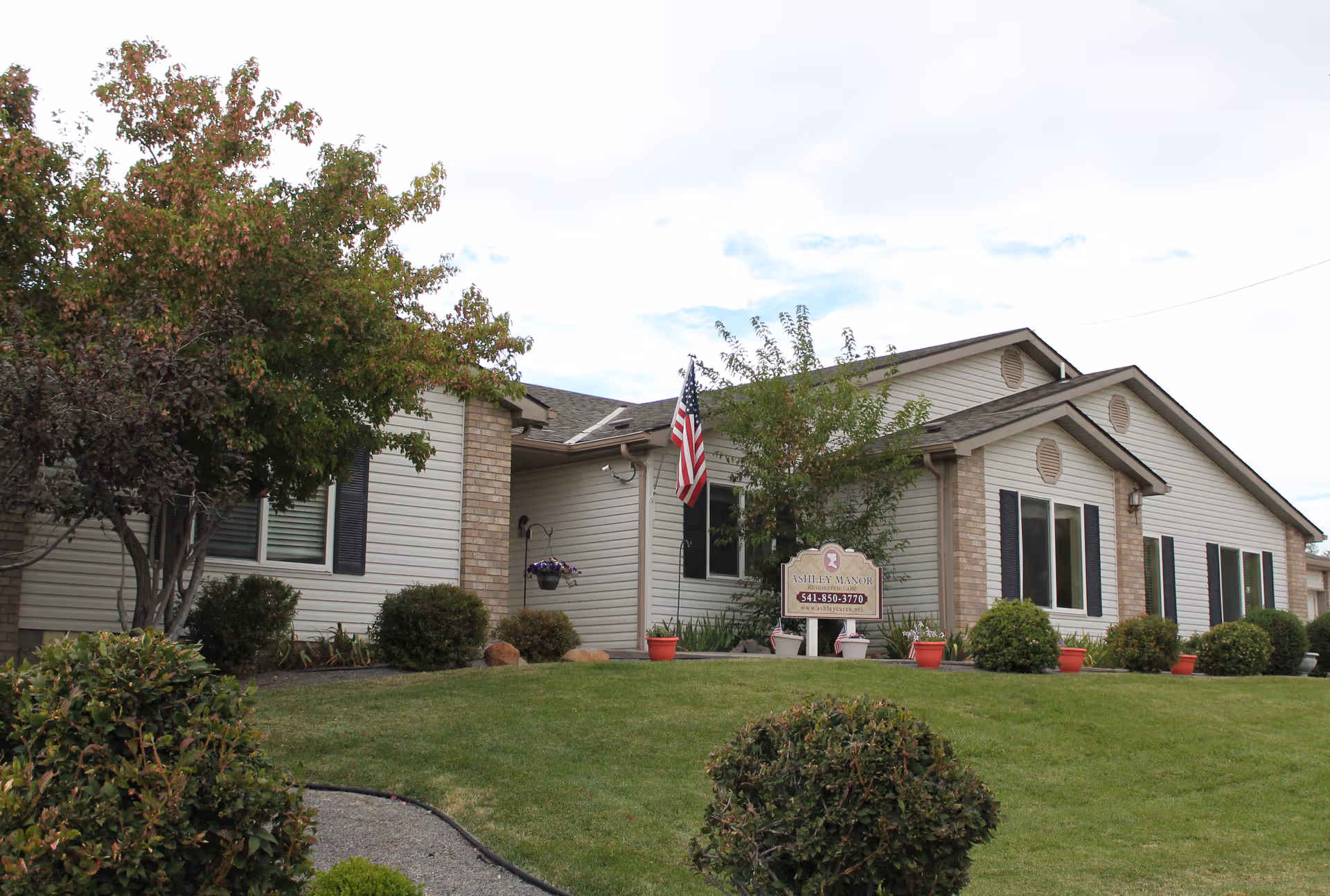 Front exterior of a single-story senior living facility with lawn, shrubs, and an American flag.