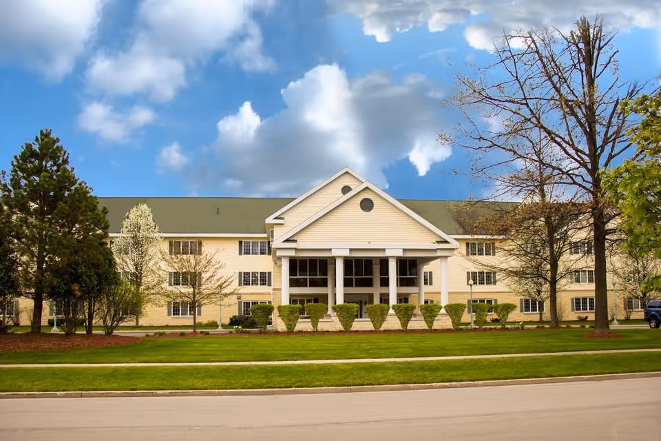 Front exterior view of a large, three-story senior living facility building with a green roof, beige siding, and a prominent entrance with white columns. The building is surrounded by neatly trimmed bushes, trees, and a well-maintained lawn under a partly cloudy blue sky.