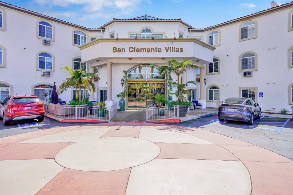 Front entrance of the San Clemente Villas building with a covered portico, potted palms, and cars parked at a circular drive.