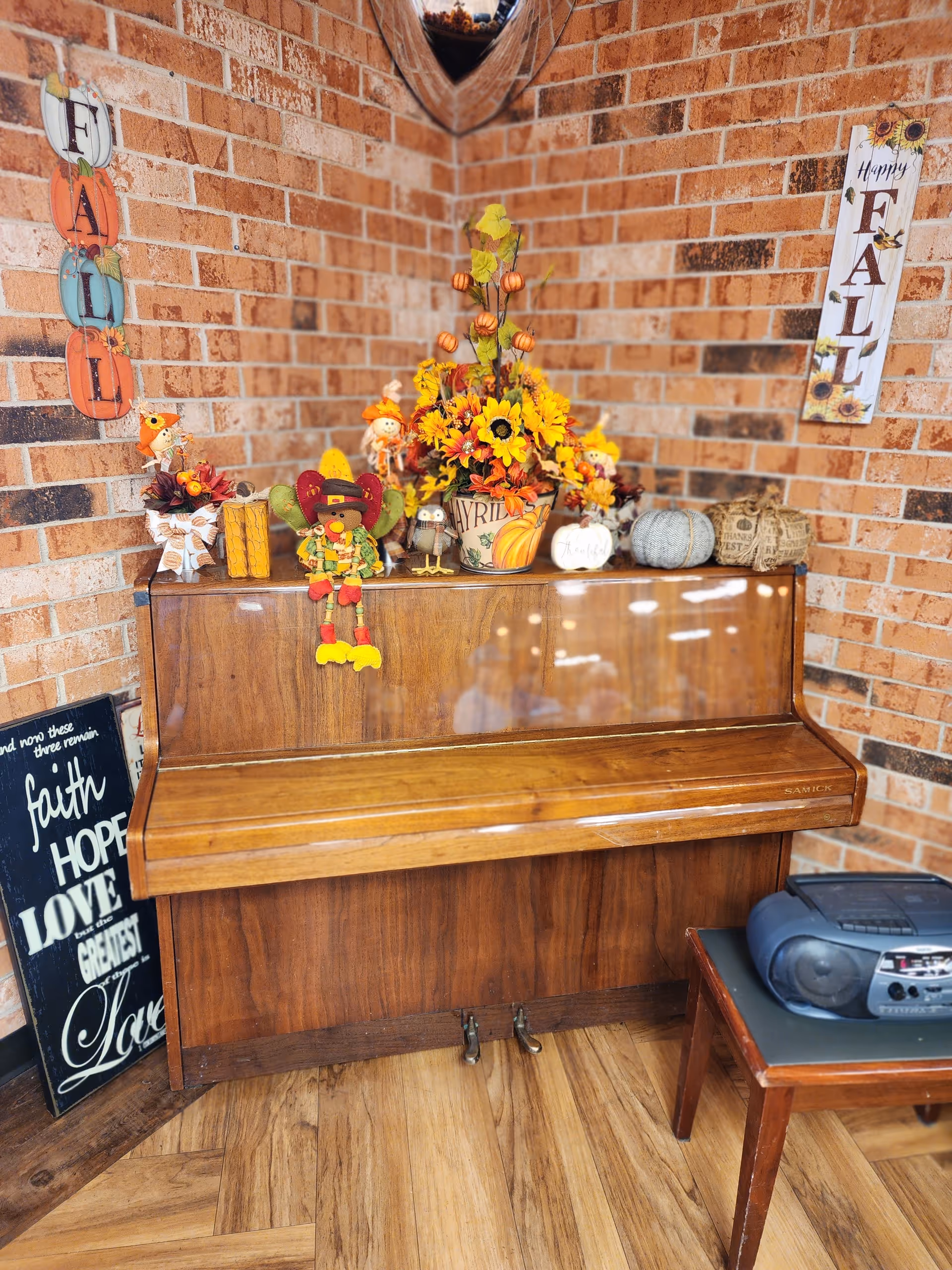 Indoor brick-walled corner with a wooden piano, autumn decorations, and a small radio.