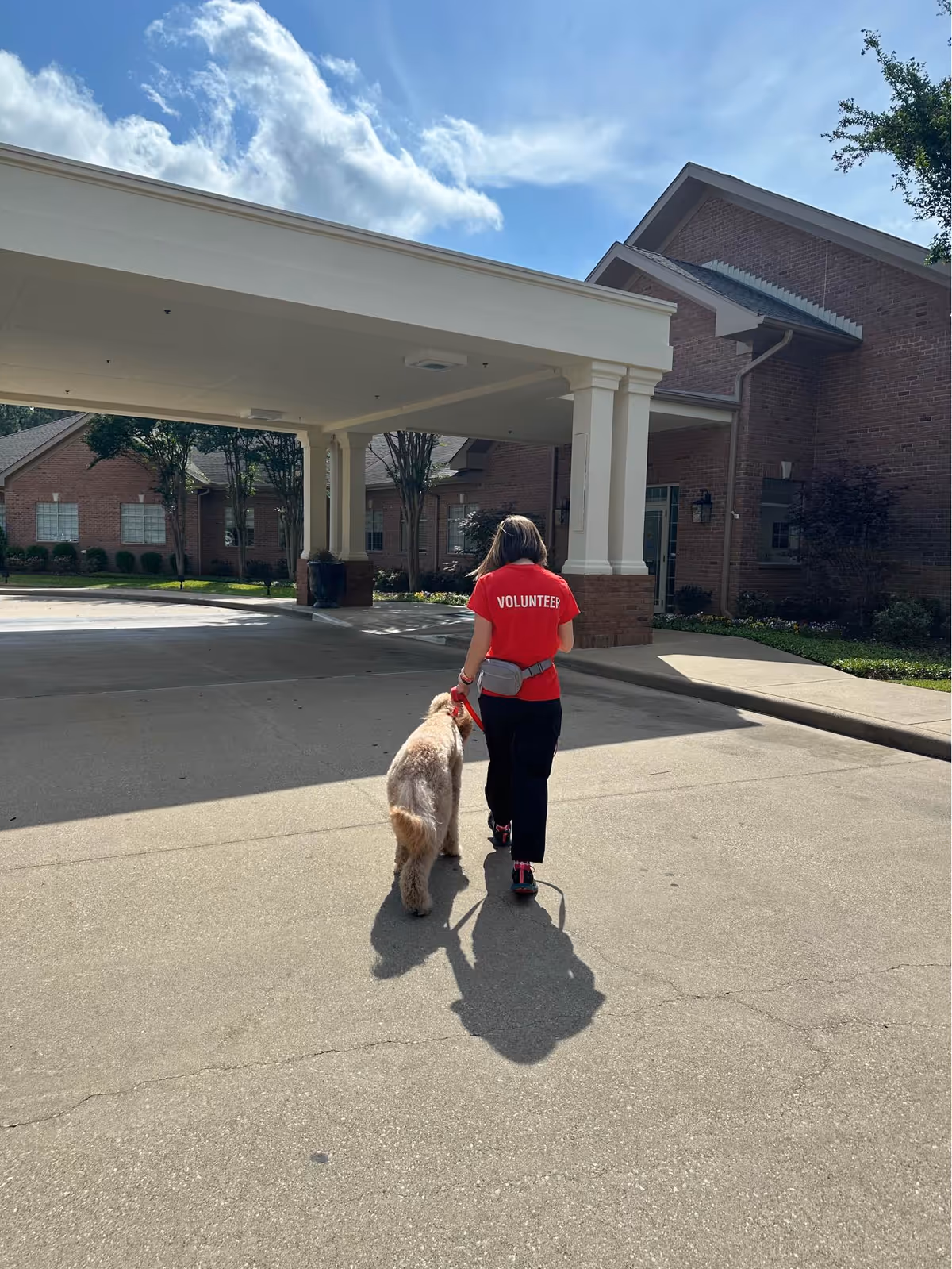 Volunteer in a red shirt walks a dog outside the hospice facility under a covered entrance