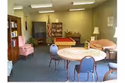 Cozy senior living lounge with a round table, chairs, sofas, bookshelf, and TV.
