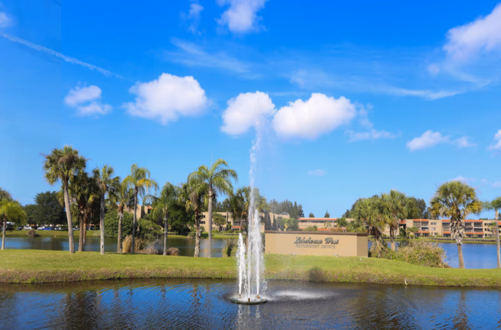 A pond with a central fountain, palm trees, and the Lakehouse West Retirement Estate buildings and sign under a bright blue sky.