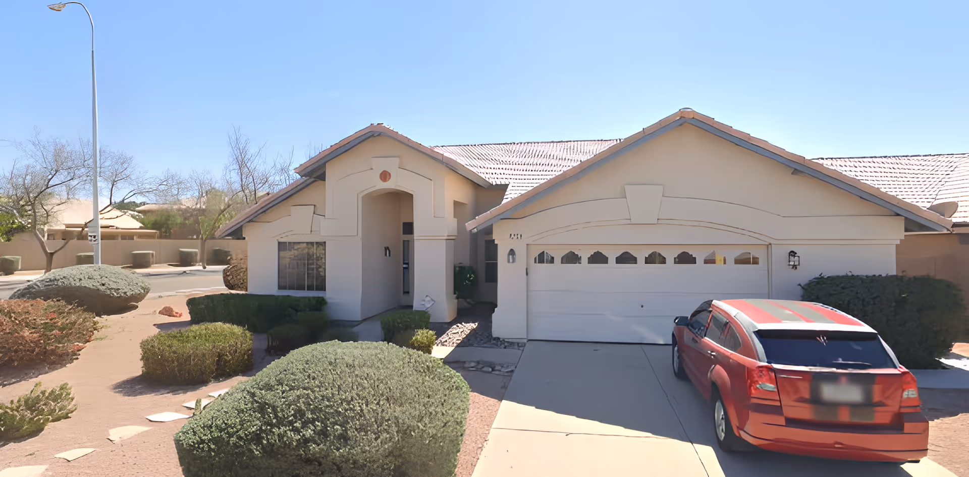 Single-story stucco house with an attached garage, desert landscaping, and a red car parked in the driveway under a clear sky.