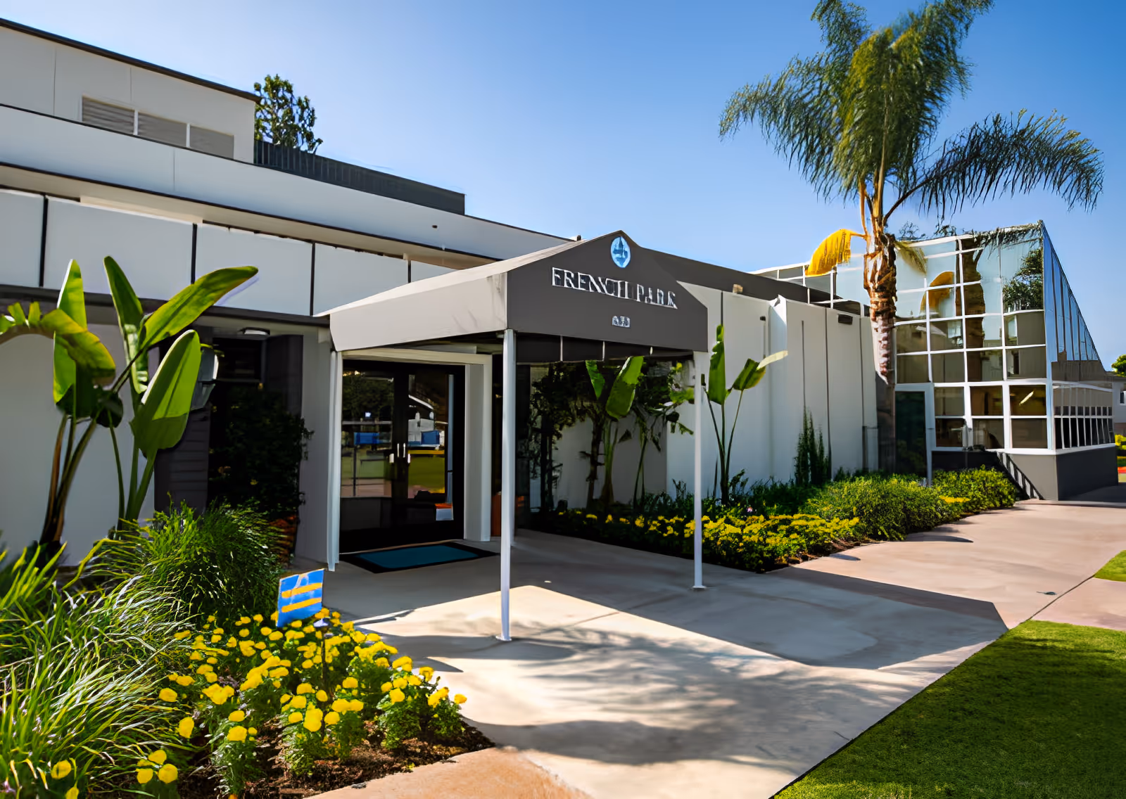 Front entrance of French Park Care Center with a covered awning, landscaped flowerbeds and palm trees.