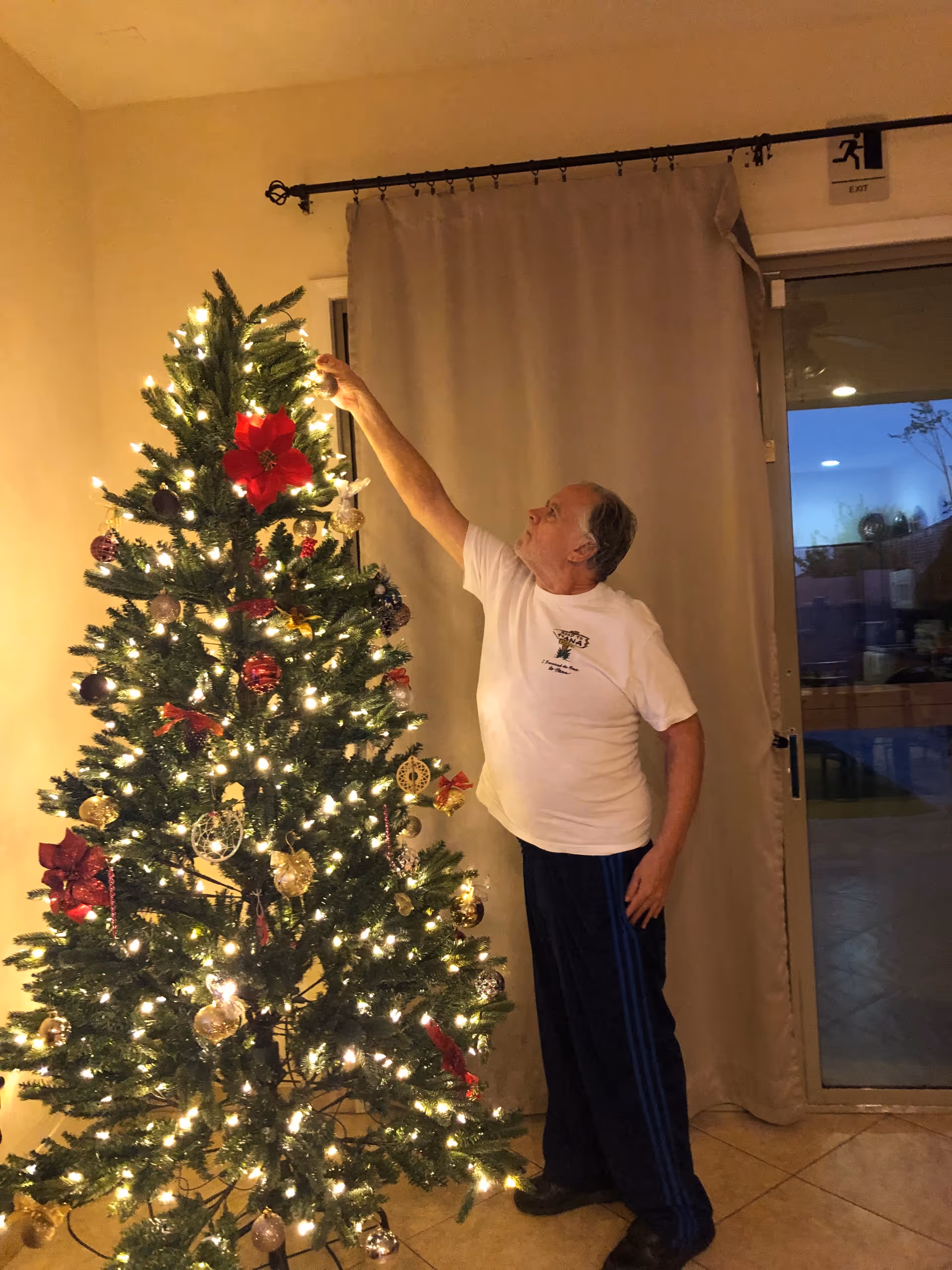 Older man decorates a Christmas tree indoors with lights.