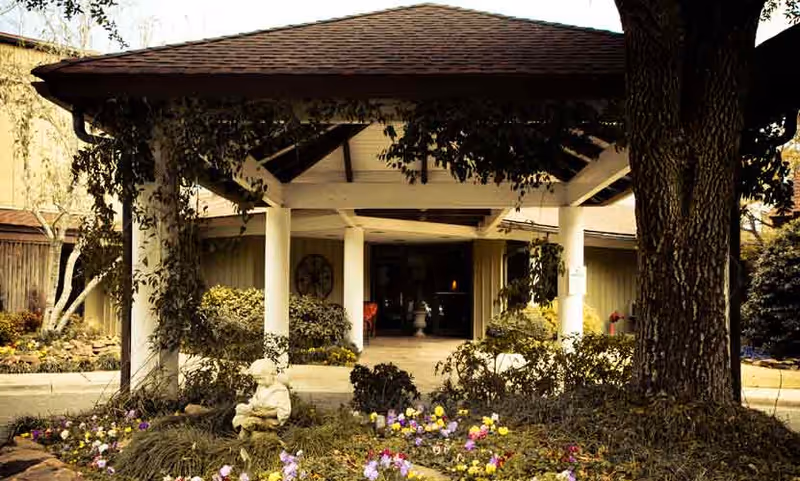 Entrance of a building with a covered porch supported by white columns, surrounded by greenery and colorful flowers. A small statue of a child is visible in the garden area near the entrance. Large trees frame the scene.