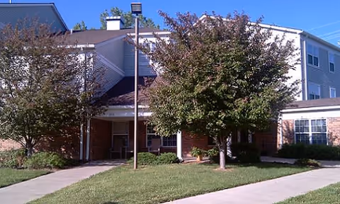 Front entrance of a multi-story senior apartment building with a covered entry, trees, and a walkway across a grassy lawn.