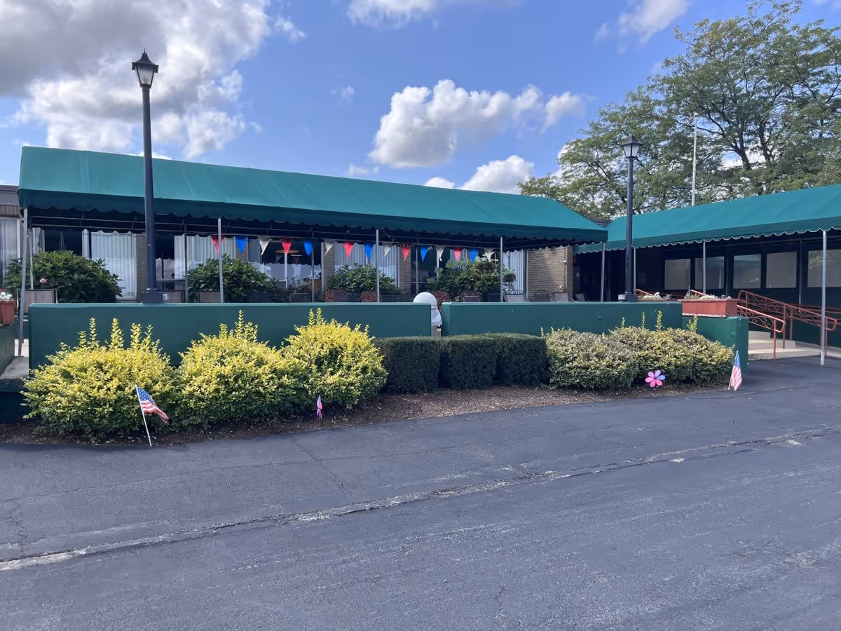 Exterior view of a building with green awnings covering a patio area. There are bushes and small American flags planted in front of the building, with a paved driveway or parking area in the foreground. The sky is partly cloudy with blue patches visible.