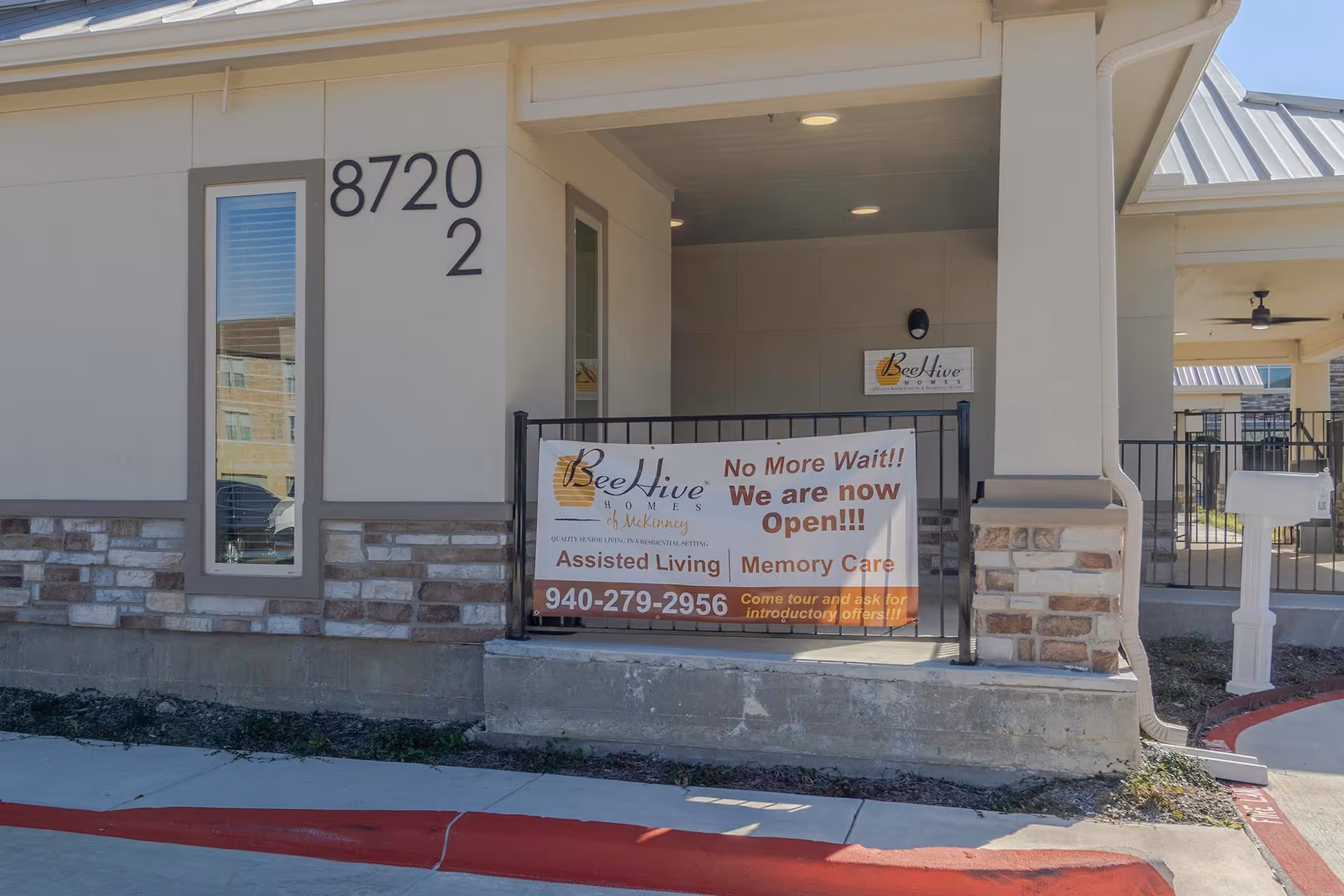 Exterior view of BeeHive Homes of McKinney showing the building entrance with a sign that reads 'No More Wait!! We are now Open!!! Assisted Living | Memory Care' along with a phone number and invitation to tour and ask for introductory offers.
