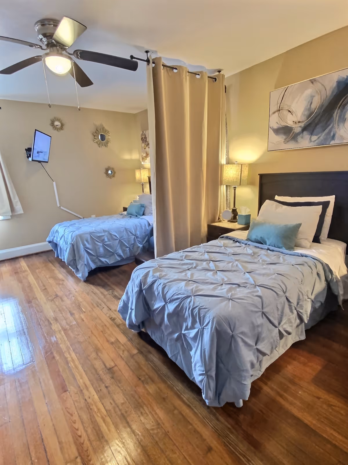 Twin beds with blue quilts separated by a curtain, wooden floors, and lamps.