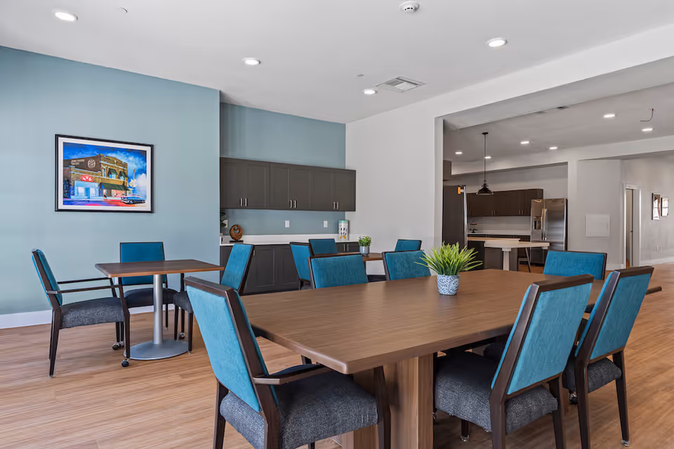 A modern dining area with blue chairs around a large wooden table and kitchenette in back.