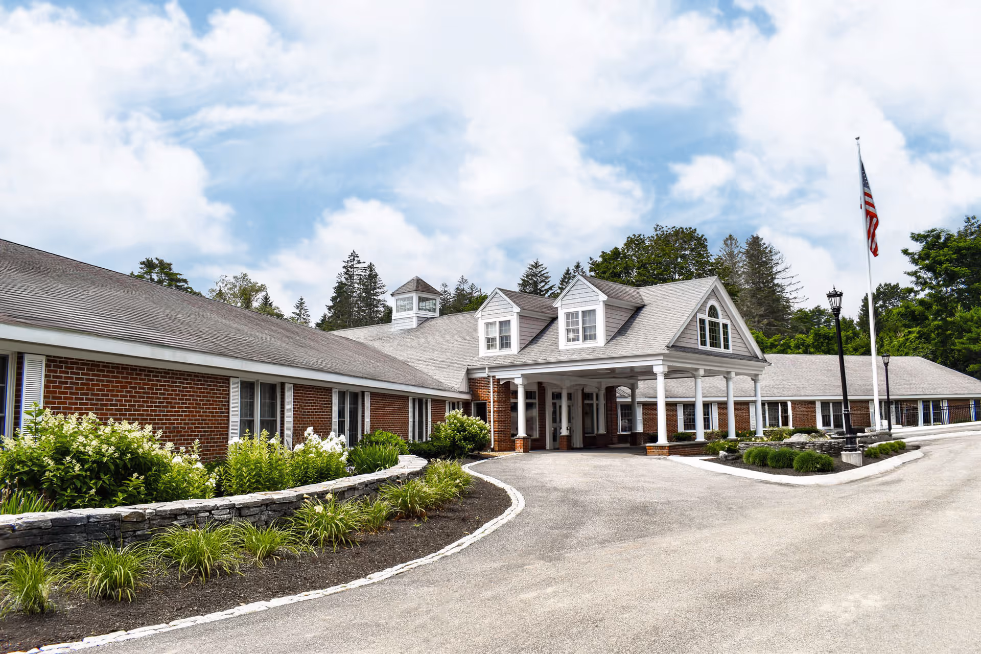 Brick senior living facility exterior with a covered entrance, white columns, and American flag.