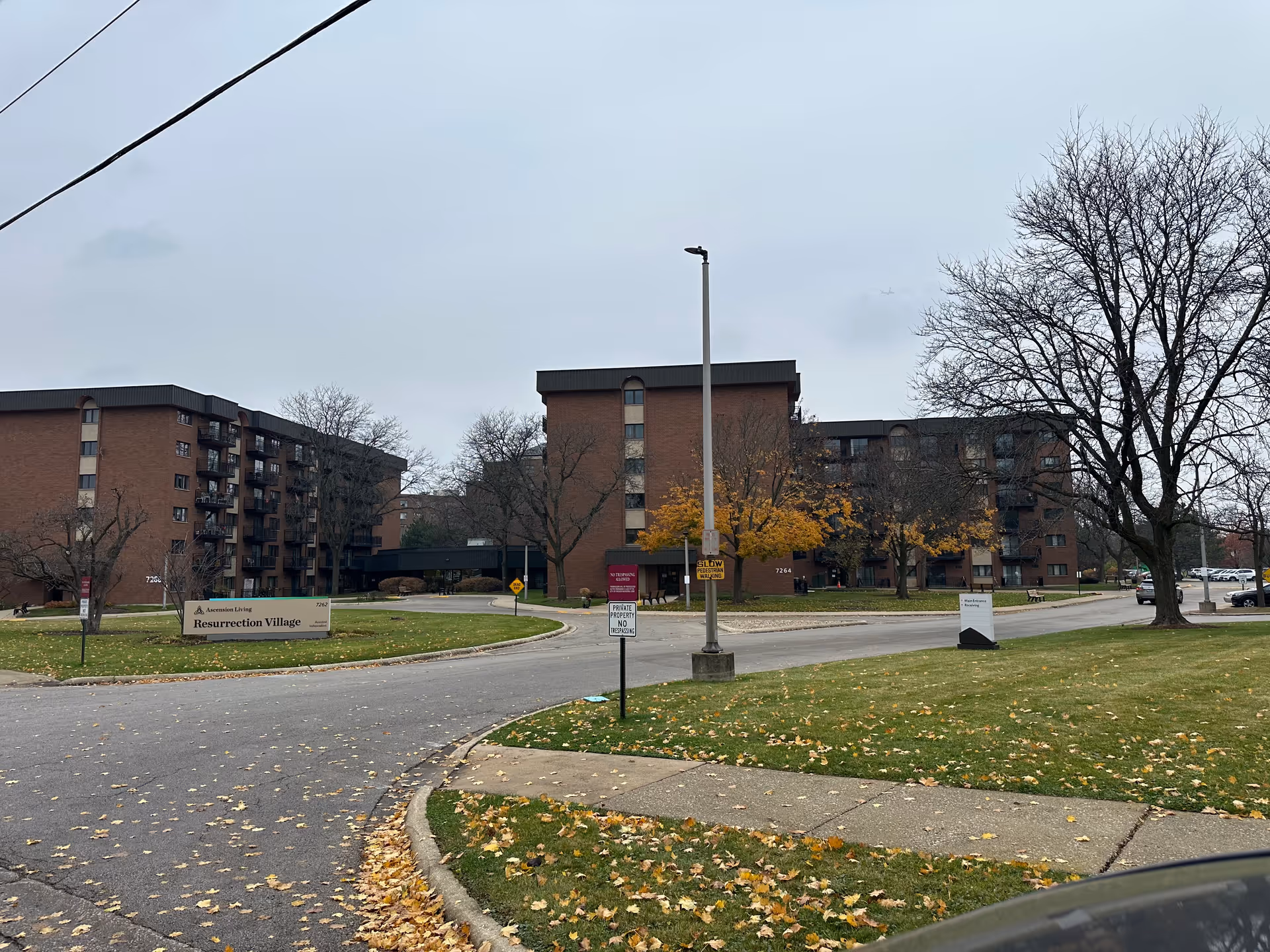 Brick multi-story residential buildings of Ascension Living Resurrection Village seen from the driveway with lawn, trees and entrance signage.