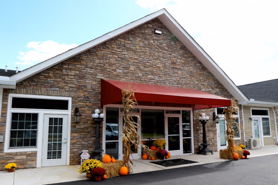Entrance of a building with a stone facade and a red awning above glass double doors. The entrance is decorated with autumn-themed items including pumpkins, hay bales, corn stalks, and potted chrysanthemums. There are also two black lamp posts with globe lights on either side of the entrance.