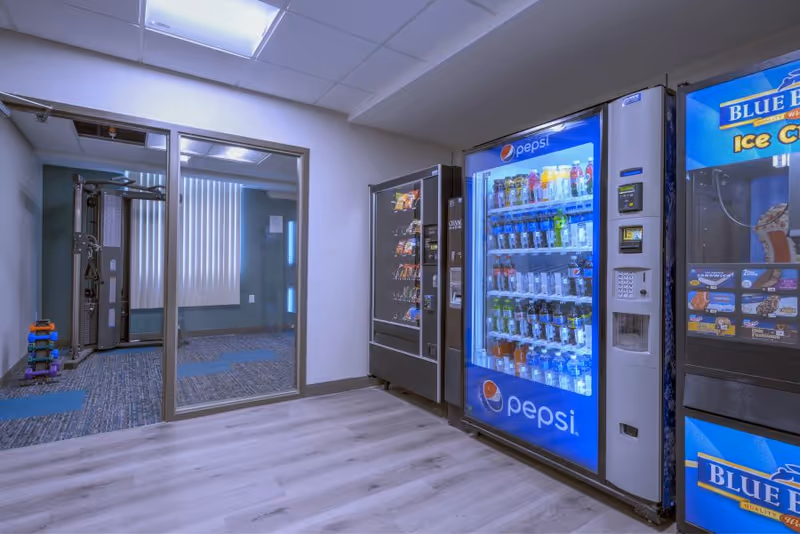 Interior area with a Pepsi drink vending machine and snack machines beside a glass-walled fitness room.