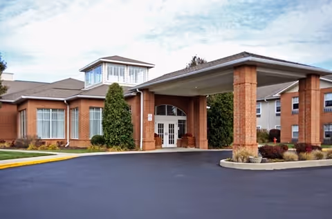 Exterior view of a senior living facility with a covered entrance supported by brick columns, large windows, and a paved driveway. The building is surrounded by some landscaping including bushes and small trees under a partly cloudy sky.
