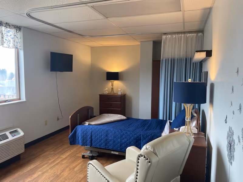Hospital-style patient bedroom with blue quilted bed, nightstands, lamps, and a chair.