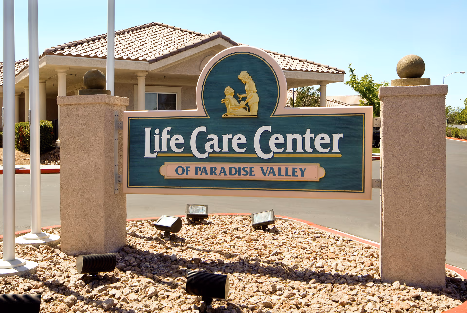 Outdoor view of a sign for Life Care Center of Paradise Valley mounted between two stone pillars with a building and driveway in the background under a clear sky.