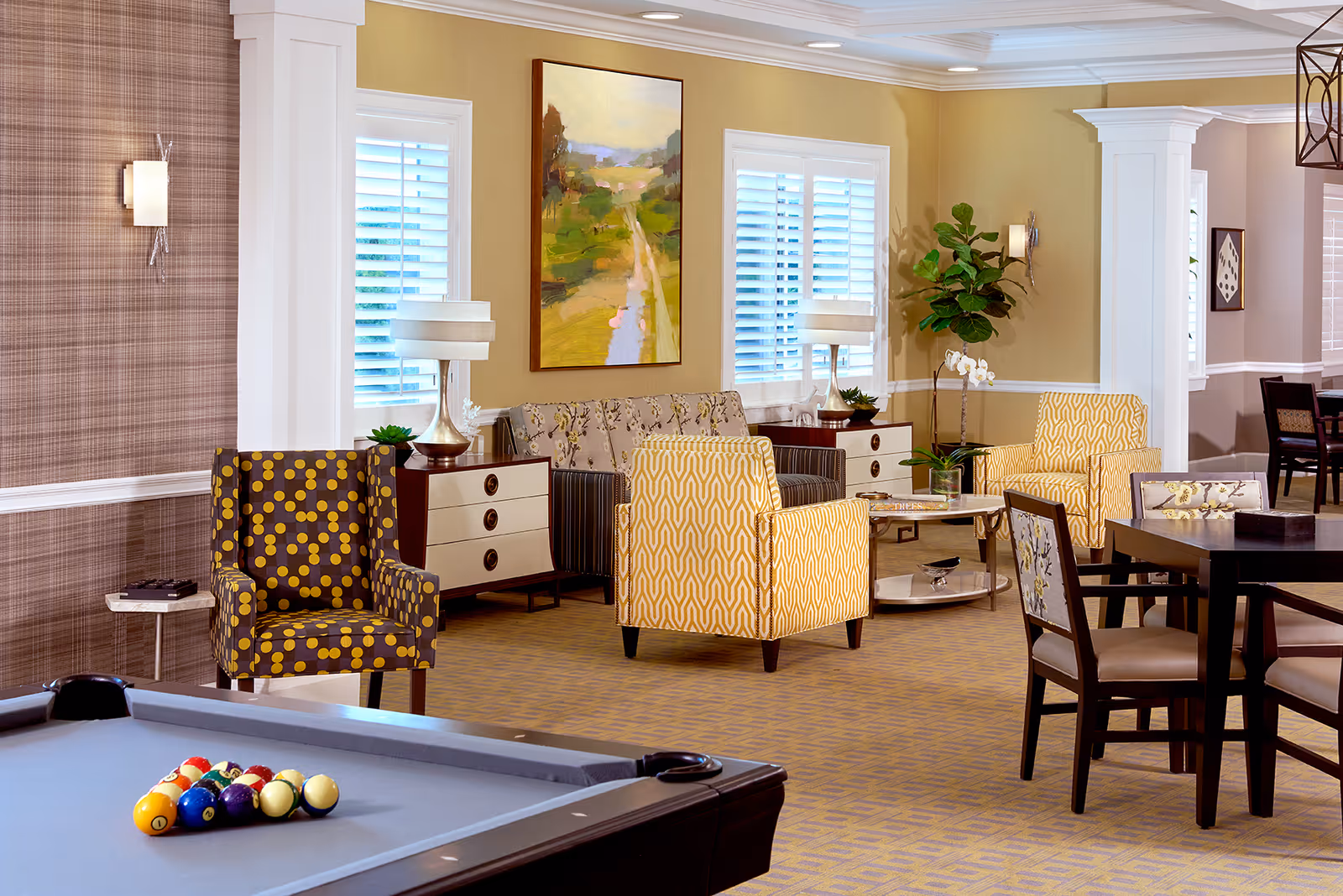 A well-lit common area in an assisted living facility featuring a pool table in the foreground, several patterned armchairs, a sofa, side tables with lamps, a coffee table with decorative items, and a dining table with chairs. The walls are decorated with framed artwork and there are windows with white shutters allowing natural light in.