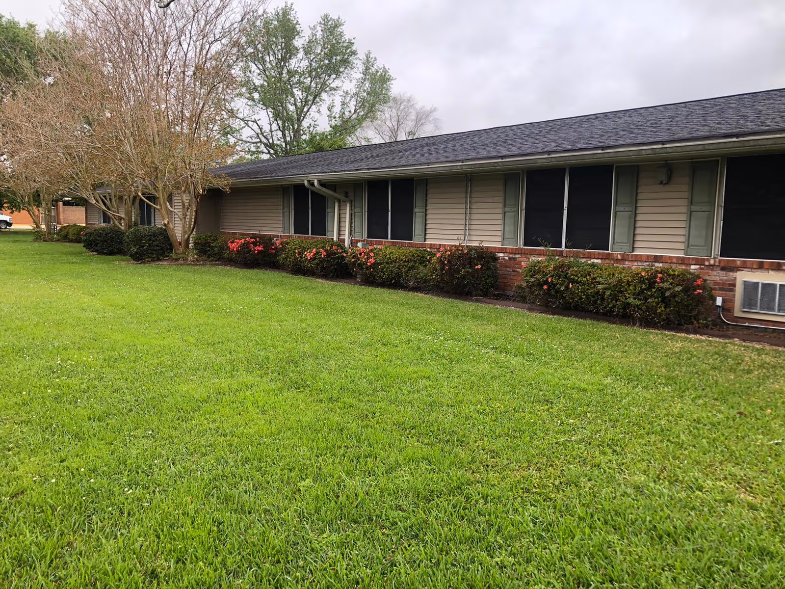 Single-story building with beige siding and green shutters, surrounded by a well-maintained lawn and bushes with pink flowers under a cloudy sky.