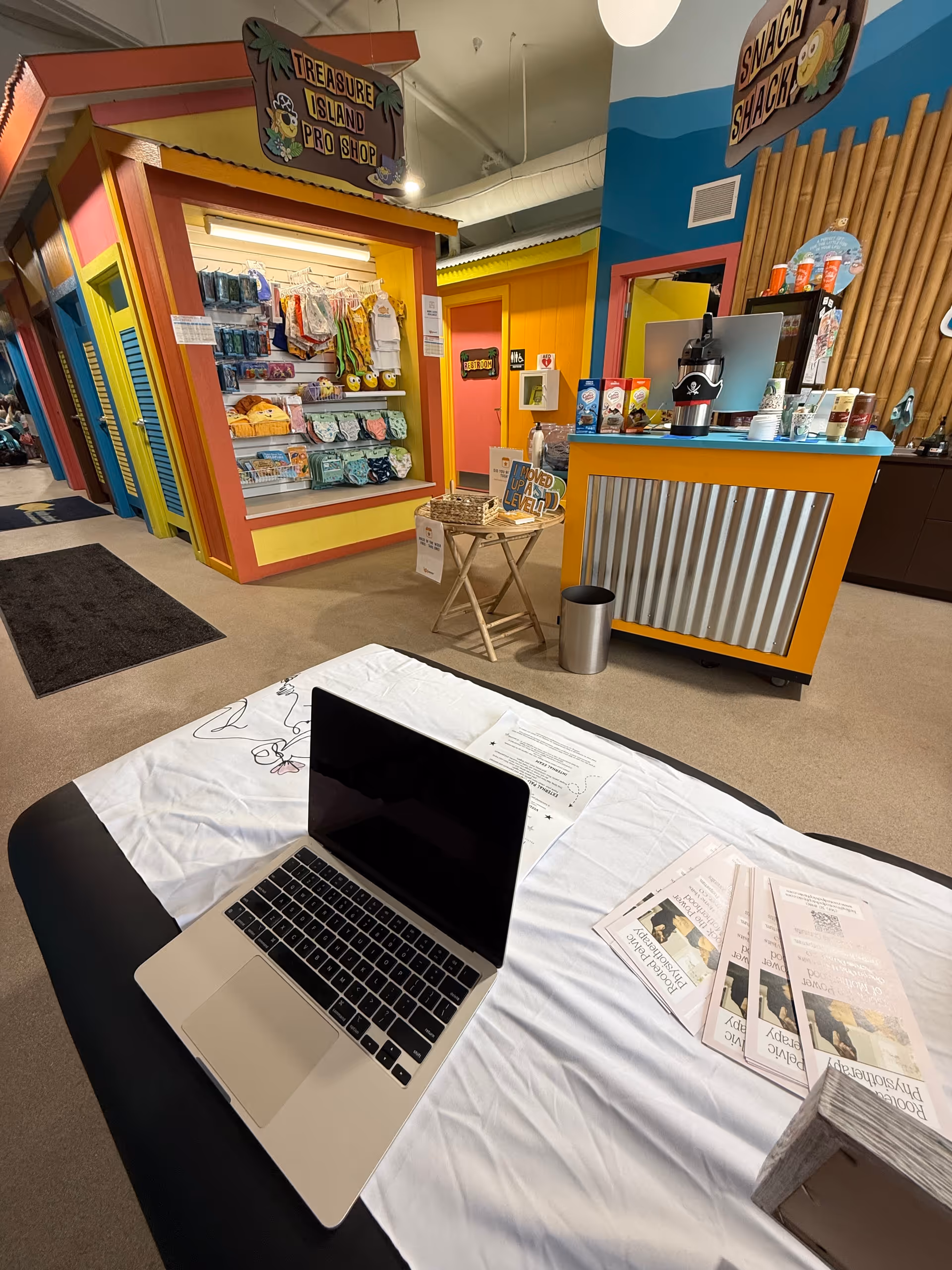 Colorful indoor space with storefronts, a laptop on a table, and brochures.