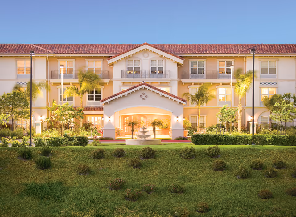 Front exterior of a three-story Mediterranean-style senior living building with a lit entrance, fountain, palm trees, and landscaped lawn.