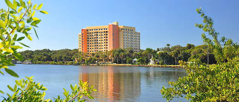 A large multi-story waterfront building with palm trees and its reflection in calm water framed by greenery.