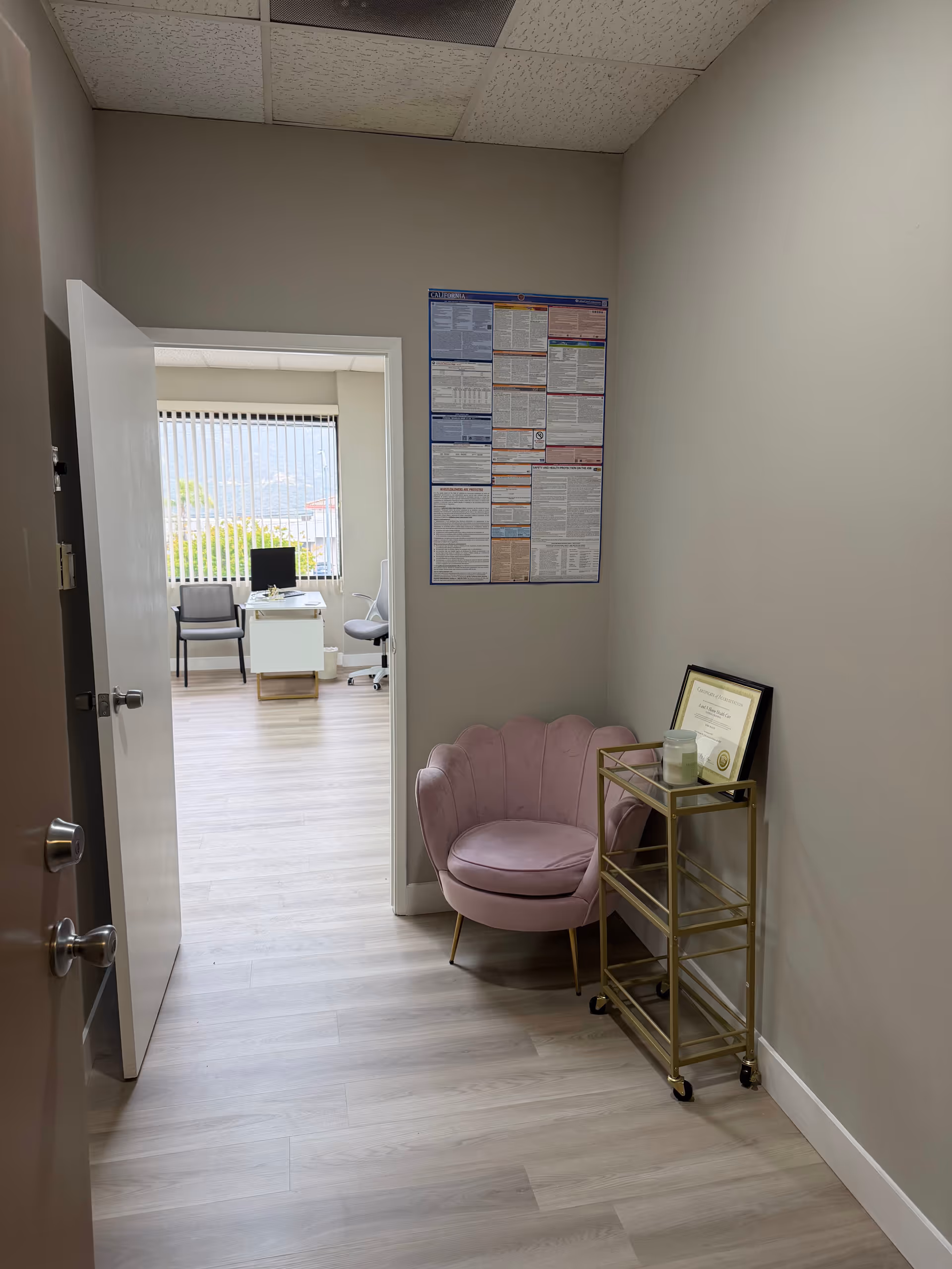 View down a hallway to an office with a pink chair, cart, and wall chart