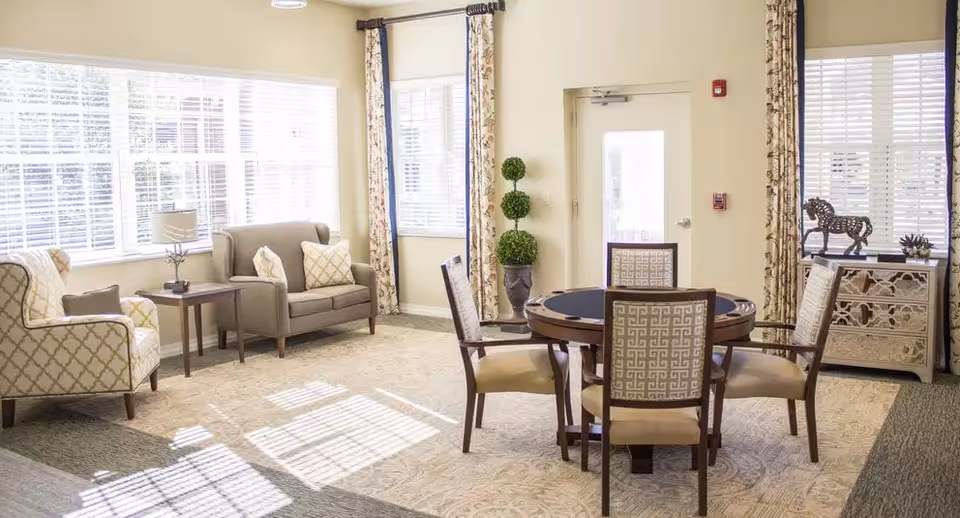 A bright and cozy living room area with large windows covered by white blinds allowing natural light to fill the space. The room features a beige loveseat with patterned cushions, an armchair with a geometric pattern, a small side table with a lamp, and a round wooden table surrounded by four upholstered chairs. There are decorative curtains, a potted topiary plant, and a cabinet with a horse sculpture and small plant on top.