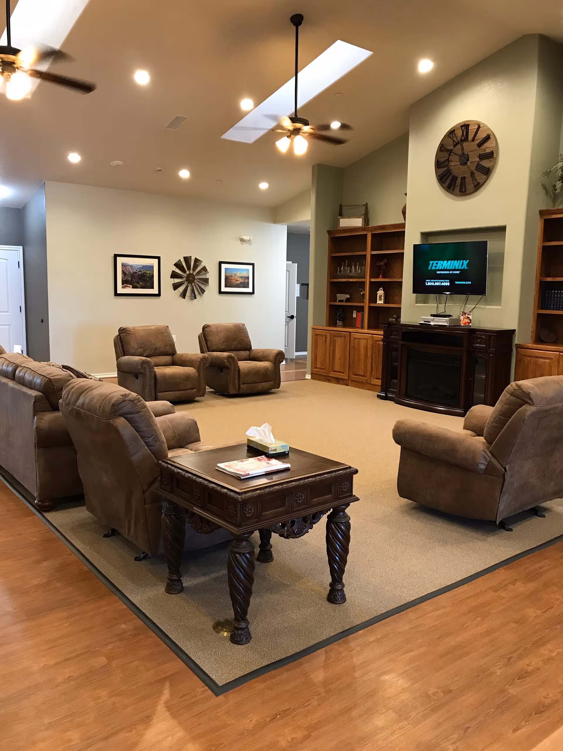 A spacious living room area with multiple brown recliner chairs and a matching sofa arranged around a wooden coffee table. The room features a large carpet over wooden flooring, ceiling fans with lights, recessed lighting, and a wall-mounted TV above a fireplace. There are built-in wooden shelves on either side of the fireplace, decorated with various items. Two framed pictures and a decorative wall clock are visible on the walls.