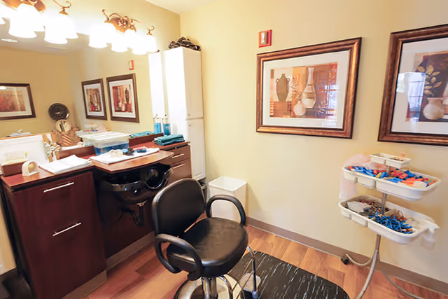 Interior of a salon area in a senior living facility with a black salon chair in front of a sink and counter with various hair care items. The room has wooden flooring, framed artwork on the wall, and a rolling cart with hair accessories and tools.