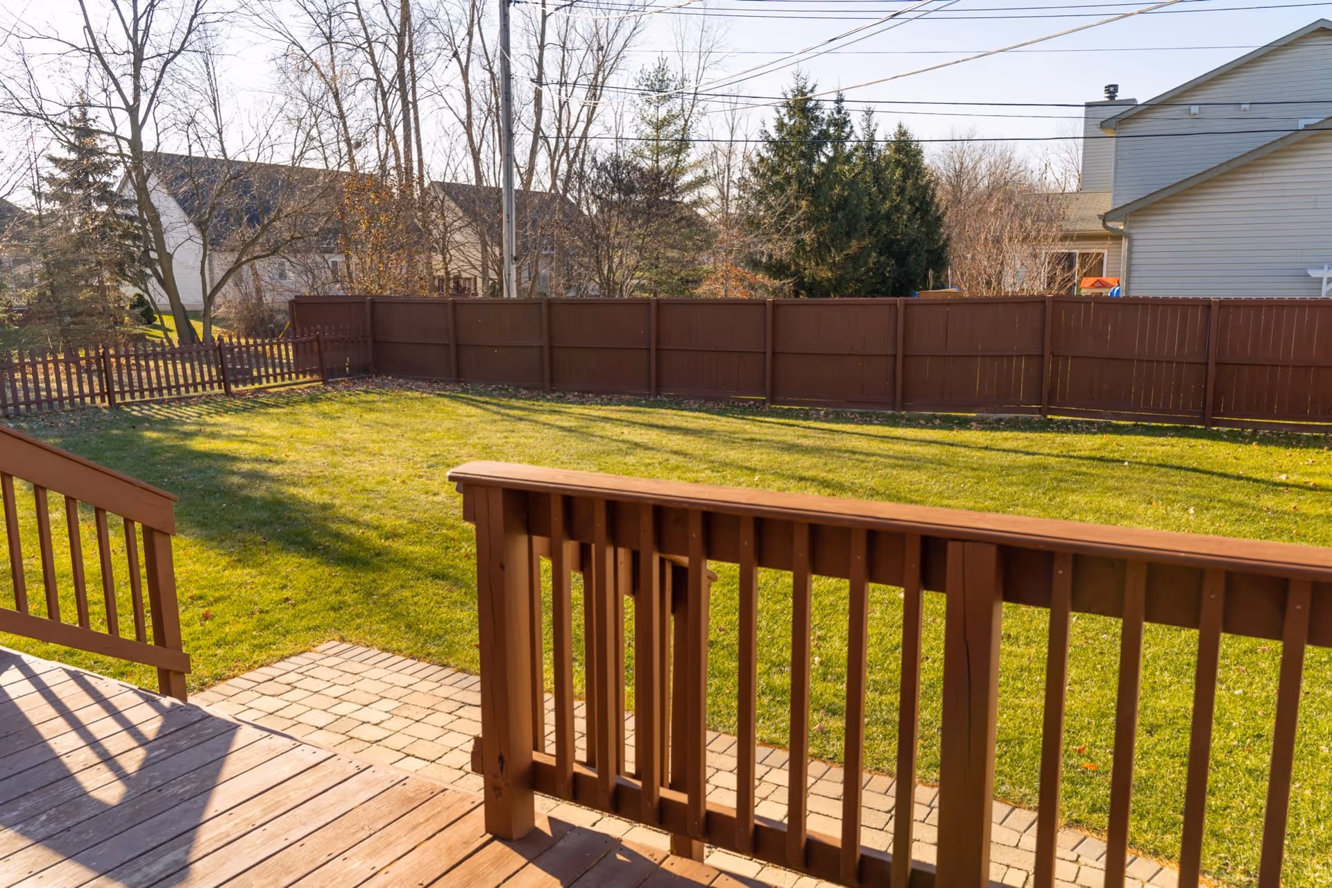 View of a fenced backyard with green grass, a wooden deck with railing in the foreground, and neighboring houses and trees in the background under a clear sky.