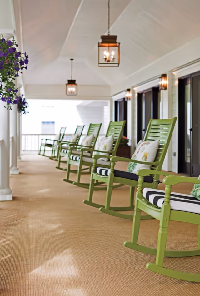 Covered porch lined with green rocking chairs with cushions and hanging lanterns.