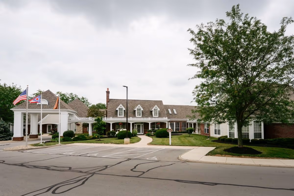 Exterior view of Sunrise of Findlay senior living facility showing a large building with a covered entrance, multiple windows, and a well-maintained lawn with trees and shrubs. Three flagpoles with flags are visible near the entrance, and there is a parking area in front.