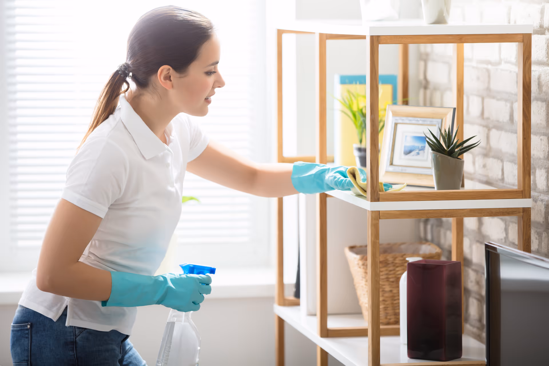 A caregiver wearing teal gloves cleans a wooden shelf in a bright interior.