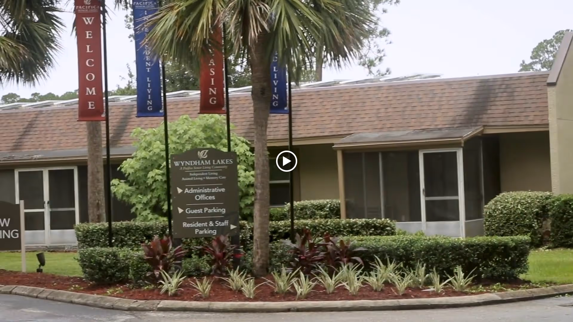 Exterior view of Wyndham Lakes senior living facility with a landscaped area featuring palm trees, shrubs, and a sign indicating directions to administrative offices, guest parking, and resident & staff parking. Several vertical banners with words like 'WELCOME', 'ASSISTED LIVING', and 'INDEPENDENT LIVING' are visible.